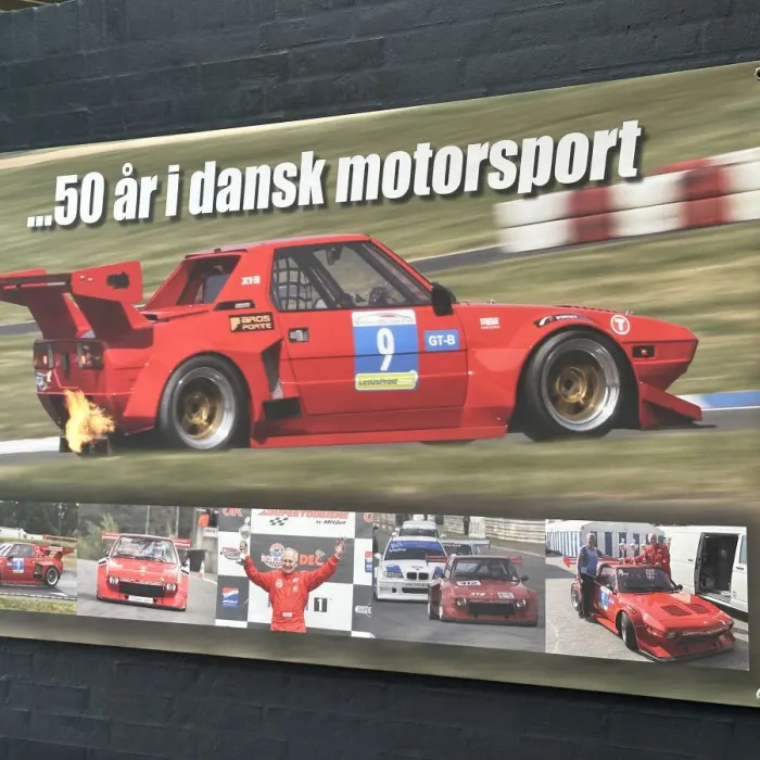 Banner ‚…50 år i dansk motorsport‘ mit Fotos eines roten Fiat X1/9 Rennwagens und seines Fahrers.